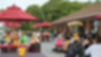 Outdoor patio with people dining at picnic tables under red umbrellas next to a brick building and lush trees.