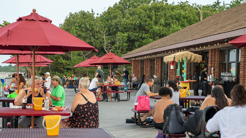 Outdoor patio with people dining at picnic tables under red umbrellas next to a brick building and lush trees.