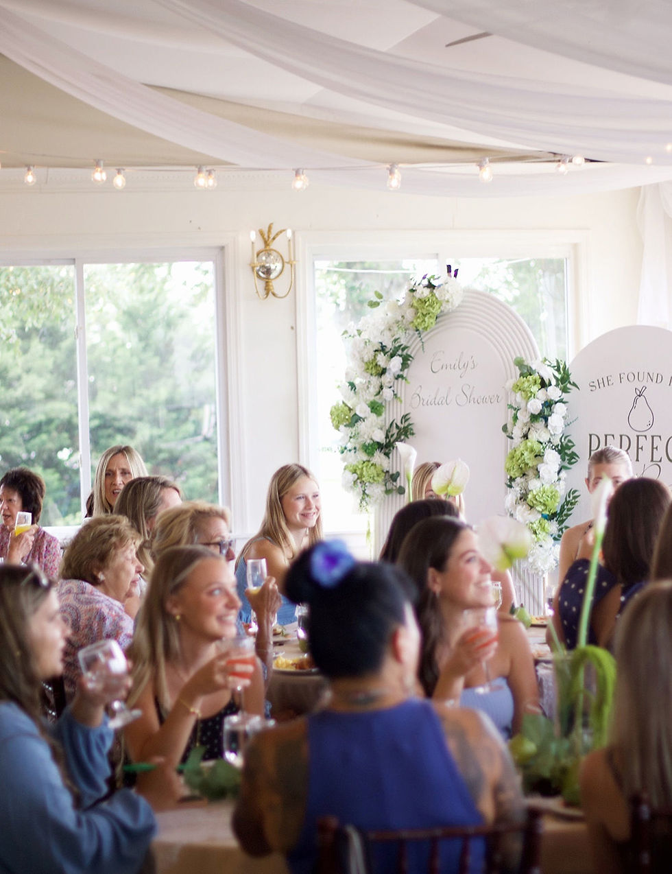 Guests smile and toast at a bridal shower celebration inside an elegantly decorated venue with white fabric drapery.