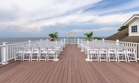 Rooftop wedding ceremony area with rows of white chairs facing a small white arch, with ocean views and palm plants on both sides.