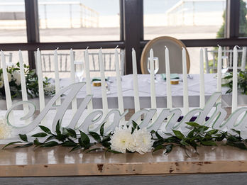A white "Alexandra" script name sign accented by white flowers, greenery, and tall taper candles.