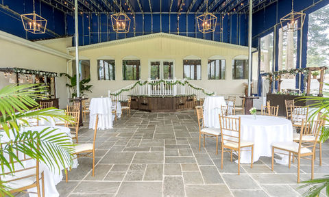 Reception space with open ceiling, stone floors, and view of a raised stage decorated with garland, surrounded by cocktail and round tables.