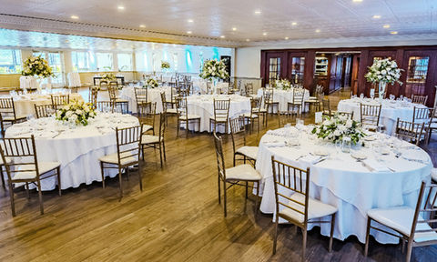 Wedding reception hall with round tables covered in white linens, gold chairs, and floral centerpieces arranged throughout the room.