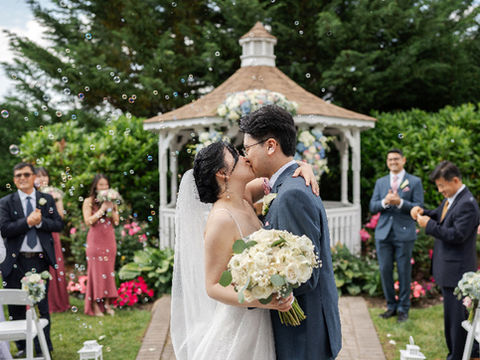 Newlyweds kissing in front of a white gazebo while guests clap and blow bubbles during an outdoor wedding ceremony.