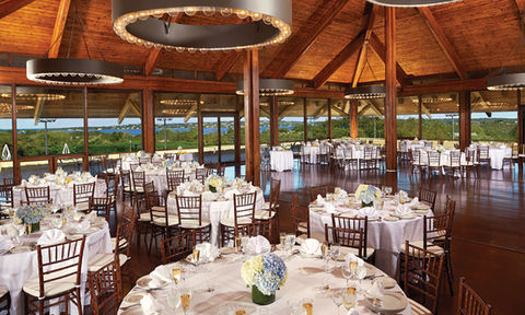 Bright reception space with panoramic windows, white linens, and waterfront views in a wood-ceiling venue.