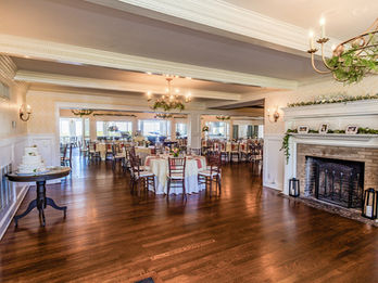 Wedding reception room with round tables, chandeliers, a white fireplace decorated with greenery, and a tiered wedding cake on a small table.