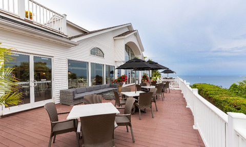 Outdoor terrace with tables, chairs, black umbrellas, and a sectional couch, overlooking the ocean beside a white building with large windows.