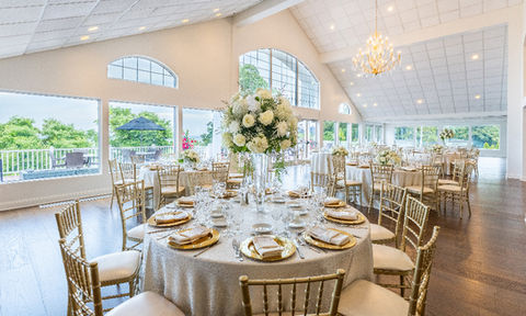 Bright wedding reception space with round tables and gold chairs arranged near large windows showing trees and outdoor umbrellas.