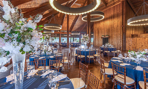 Elegant wedding reception setup with navy tablecloths, white floral centerpieces, and circular chandeliers in a rustic wood beamed hall.
