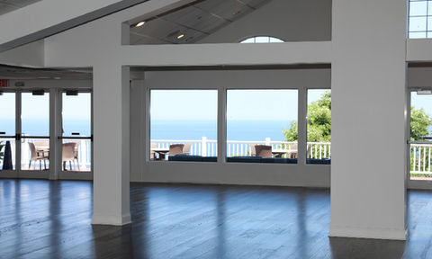 Interior view of a ballroom looking toward large windows and a patio with chairs, offering a scenic ocean view and surrounding greenery.