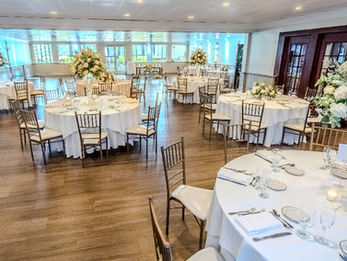 Wedding reception hall with round tables covered in white linens, floral centerpieces, gold chairs, and place settings for guests.