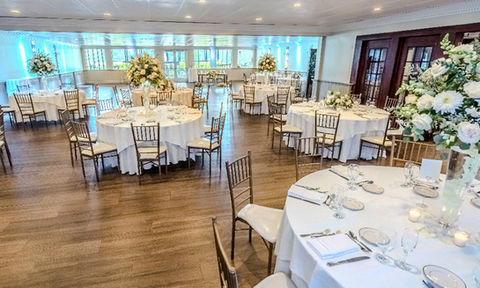 Wedding reception hall with round tables covered in white linens, floral centerpieces, gold chairs, and place settings for guests.