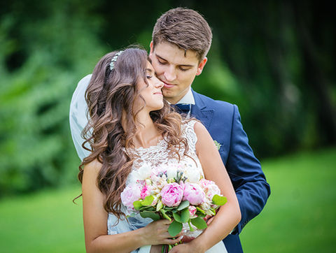 Bride holding a bouquet of pink flowers while the groom embraces her from behind in a grassy outdoor setting.