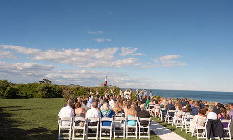 Guests seated at an outdoor wedding ceremony facing the couple and officiant, with a lighthouse and ocean in the background under a blue sky.