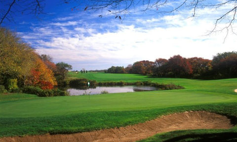 Picturesque golf green beside a pond, surrounded by colorful fall foliage and open skies.