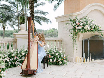 Harpist in a light blue dress playing beside a grand stone fireplace decorated with lush floral arrangements.