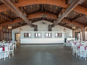 Indoor reception space with white chairs, round tables, exposed wood beams, and string lights hanging from the ceiling.