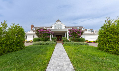 Front view of a coastal-style building facing the ocean, with manicured green lawns, sailboats on the water, and a symmetrical roofline.