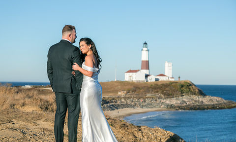 Bride and groom standing on a cliffside path with a view of the ocean and nearby lighthouse in the background.