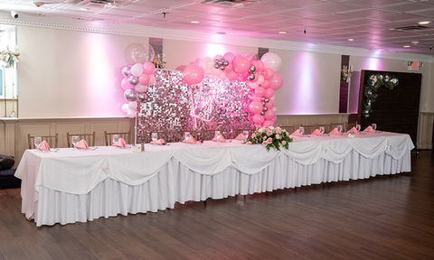 Long banquet table with pink napkins set in front of a silver sequin wall and pink balloon arch.