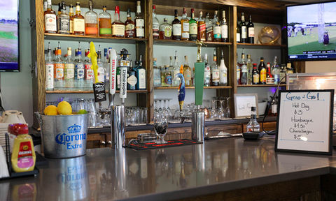 Bar counter with beer taps, a bucket of lemons, a liquor display on wooden shelves, and a small sign listing hot dog and burger prices.