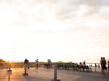 Guests gather and chat on a wooden boardwalk during sunset, overlooking the beach and ocean.