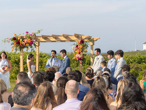 Bride and groom standing under a wooden floral arch during their outdoor wedding ceremony, with guests seated and a lighthouse visible in the background.