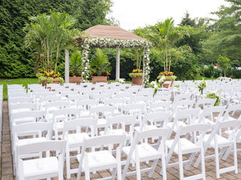 Outdoor wedding ceremony setup with rows of white chairs facing a flower-covered gazebo surrounded by greenery.