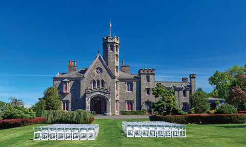 Rows of white chairs set for an outdoor ceremony on a green lawn before a majestic stone castle.