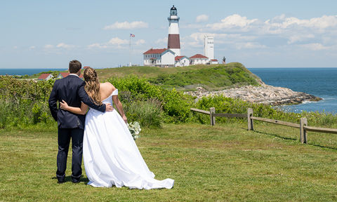 Bride and groom embracing while gazing toward a coastal lighthouse on a sunny wedding day.