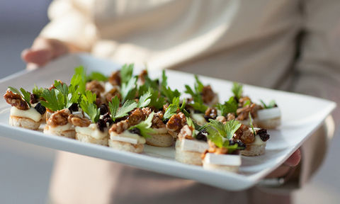 Tray of bite-sized appetizers topped with cheese, walnuts, dried fruit, and fresh parsley on round bread slices.