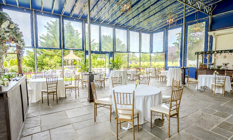 Garden-view reception space with stone tile floors, round and cocktail tables with white linens, gold chairs, and floor-to-ceiling windows overlooking a gazebo and garden.