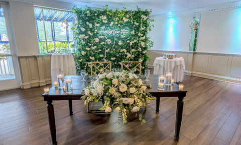 Wedding sweetheart table with floral centerpiece, glass candles, and a green flower wall in the background with a neon sign reading 'Happily Ever After'.