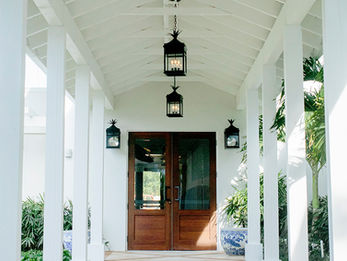 Covered walkway leading to a double wooden door entrance, surrounded by white columns and tropical plants.