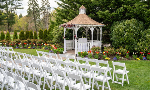 Outdoor wedding ceremony setup with rows of white chairs facing a white gazebo, surrounded by colorful tulips and greenery.