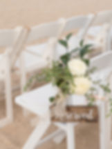 White folding wedding chair on a beach decorated with a wooden "Reserved" sign and a bouquet of white roses and greenery.