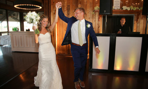 Joyful bride and groom entering their wedding reception, dancing under wooden beams and modern ring chandeliers.