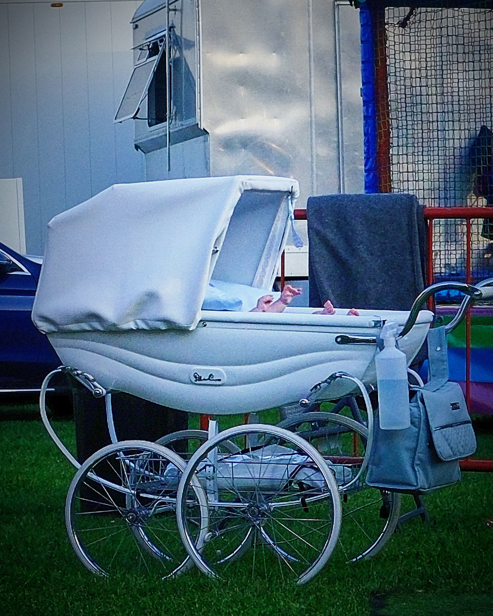 Vintage white pram at a fairground with only a baby's hands visible above the side of the carriage.