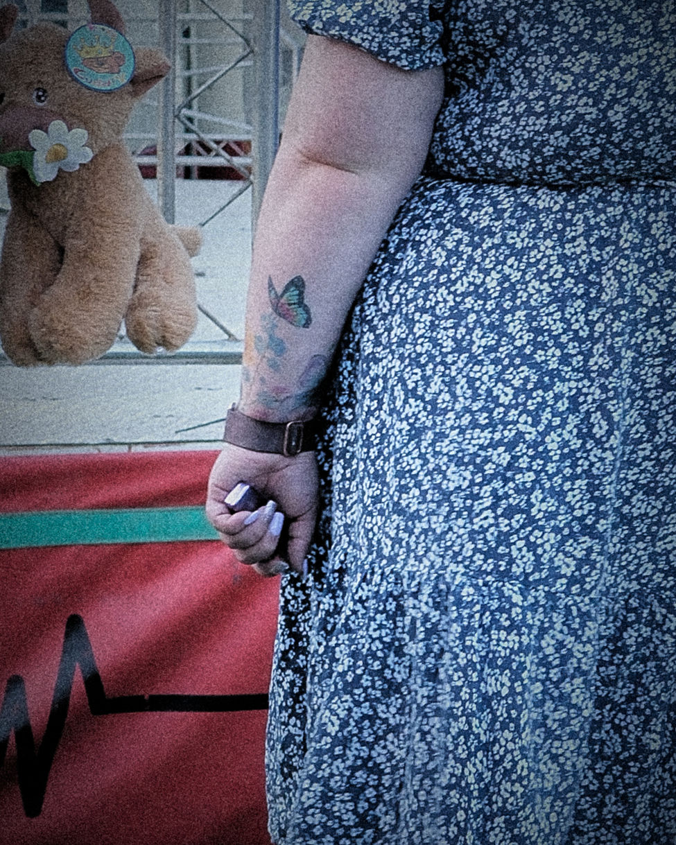 Tattooed arm holding a vape beside a fairground teddy bear with a painted heartbeat graphic in the background.