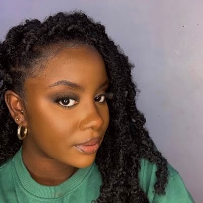 Kia wears a green jumper and poses infront of a grey wall. 