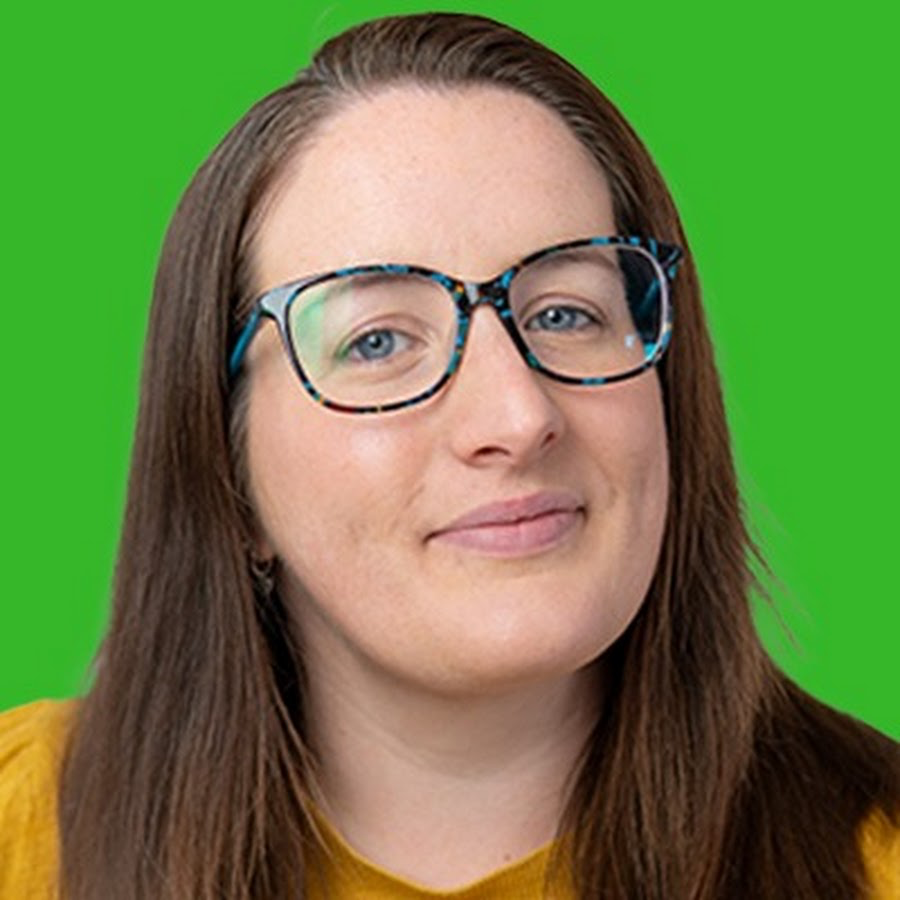Charlotte wears large blue glasses as she smiles to the camera. She poses in front of a green background. 