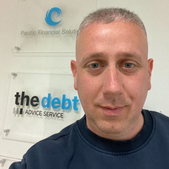 Aaron poses in front of a wall mounted 'Debt Advice Service' wall plaque. He wears a navy sweatshirt.