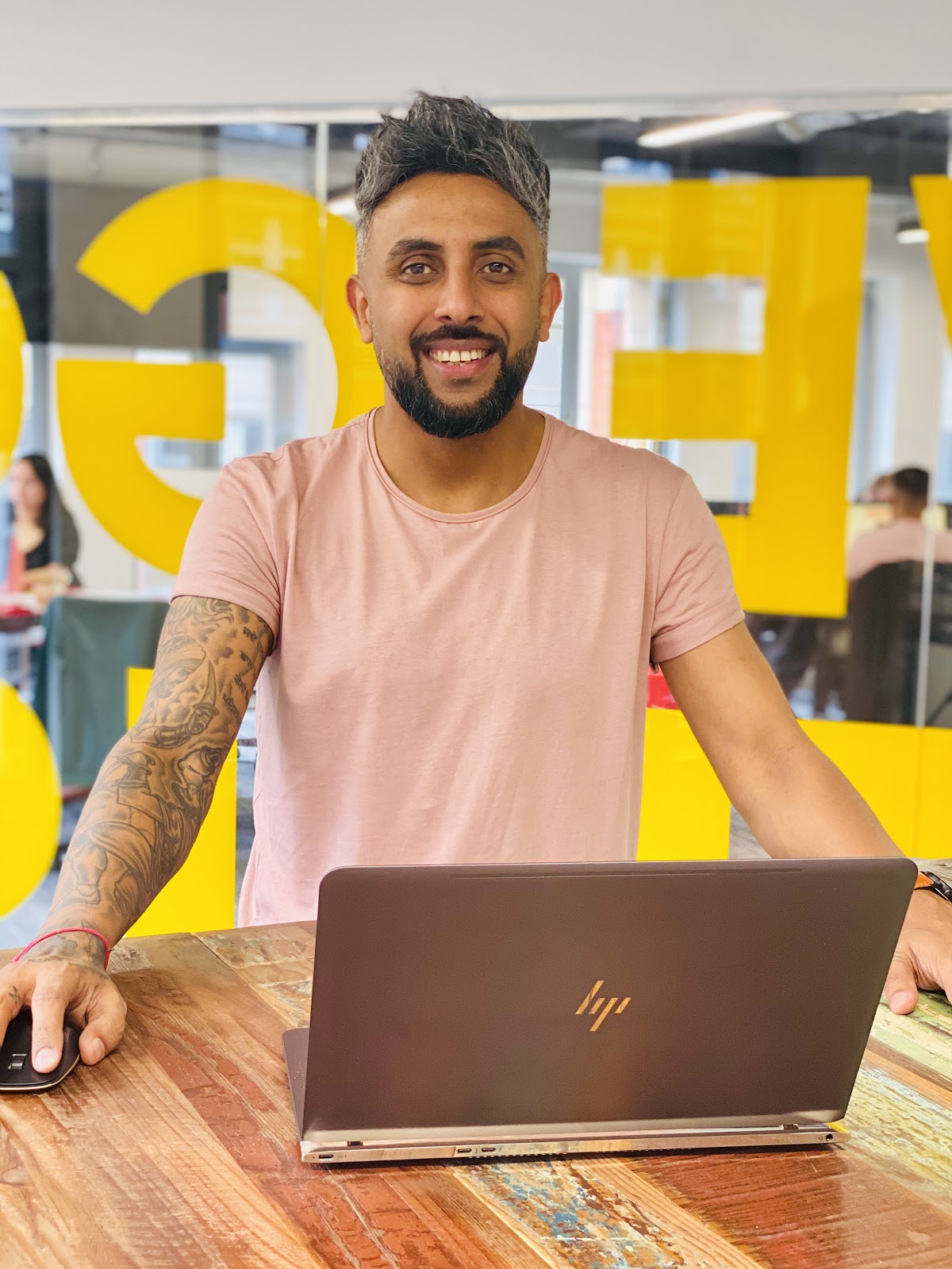 Harjit poses infront of a desk in the Freeze Debt office. He wears a pale pink t-shirt and smiles to the camera. 