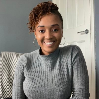 Diandra is sat in her living room, wearing a grey high neck jumper. She smiles to the camera.