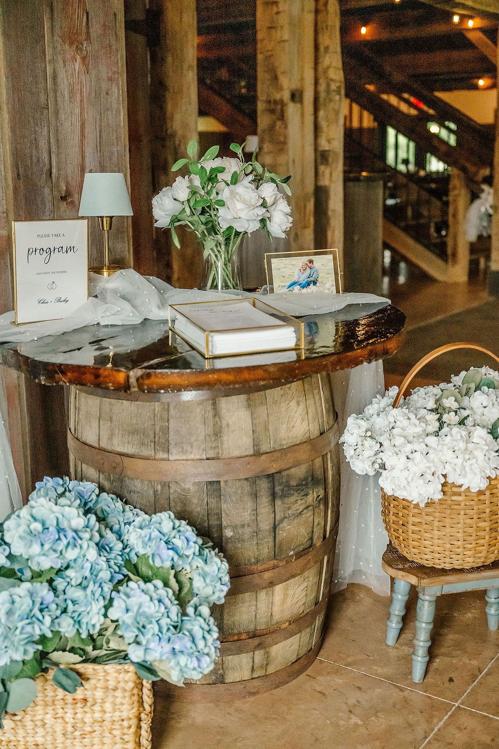 Columbus Wedding Venue near me. Rustic wooden barrel table with a program sign, flowers, and photo frame on top. Baskets of hydrangeas create a charming, cozy setting.