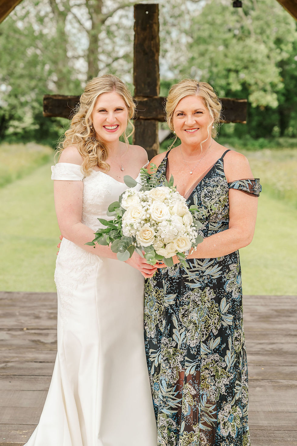 Barn Wedding Venues in Ohio. Two smiling women in dresses pose with a white rose bouquet outdoors. A wooden cross and lush green trees are in the background.
