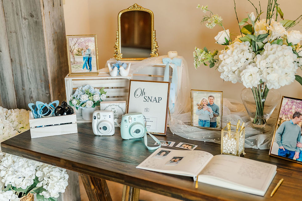 Barn Wedding Venues in Ohio. Table with photos, white flowers, cameras, and guestbook. "Oh Snap" sign stands out. Rustic wood wall backdrop, cheerful mood.