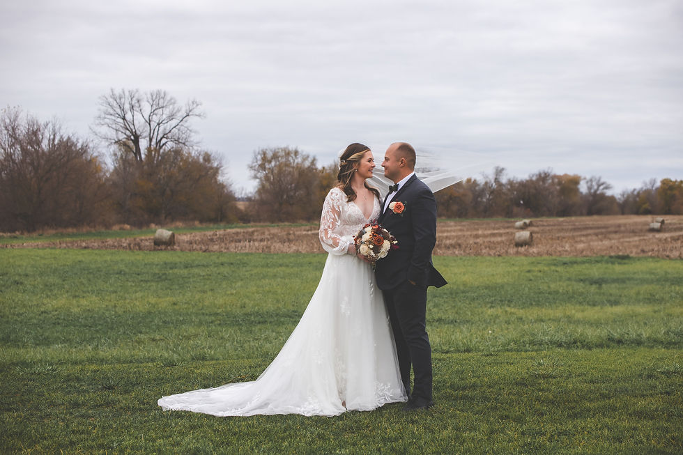 Barn Wedding Venues in Ohio. Bride and groom pose lovingly in a field, surrounded by hay bales and trees. Bride in white gown, groom in dark suit with orange boutonniere. Sky is overcast.