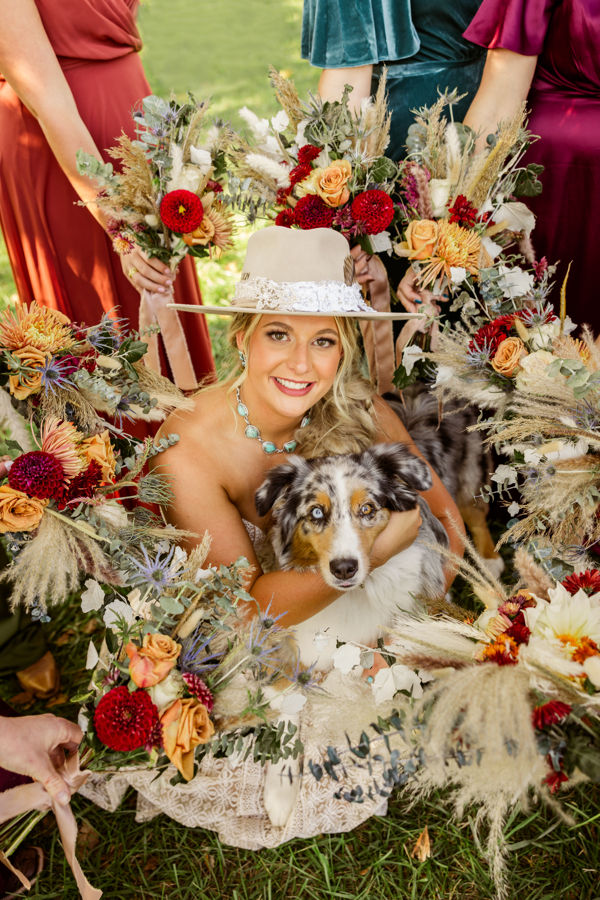 Rustic Columbus wedding venues Smiling woman in a hat holds a dog, surrounded by friends with colorful floral bouquets. Outdoors on grass, joyful and vibrant scene.