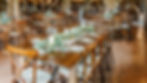 Barn Wedding Venue Ohio. Rustic dining setting with wooden tables arranged in a cozy, warmly lit venue. Tables feature elegant greenery, candles, and set plates.